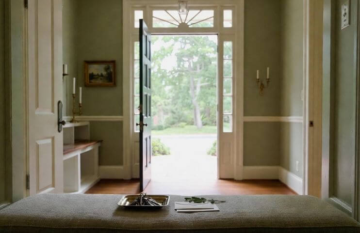 Elegant Wilmington-area home entryway with an open front door letting in soft morning light; on a sage-green bench sits a brass tray holding house keys, a welcome envelope, and a single fresh sage leaf – a quiet invitation to begin simplifying your life with Sage Lifestyle Management.